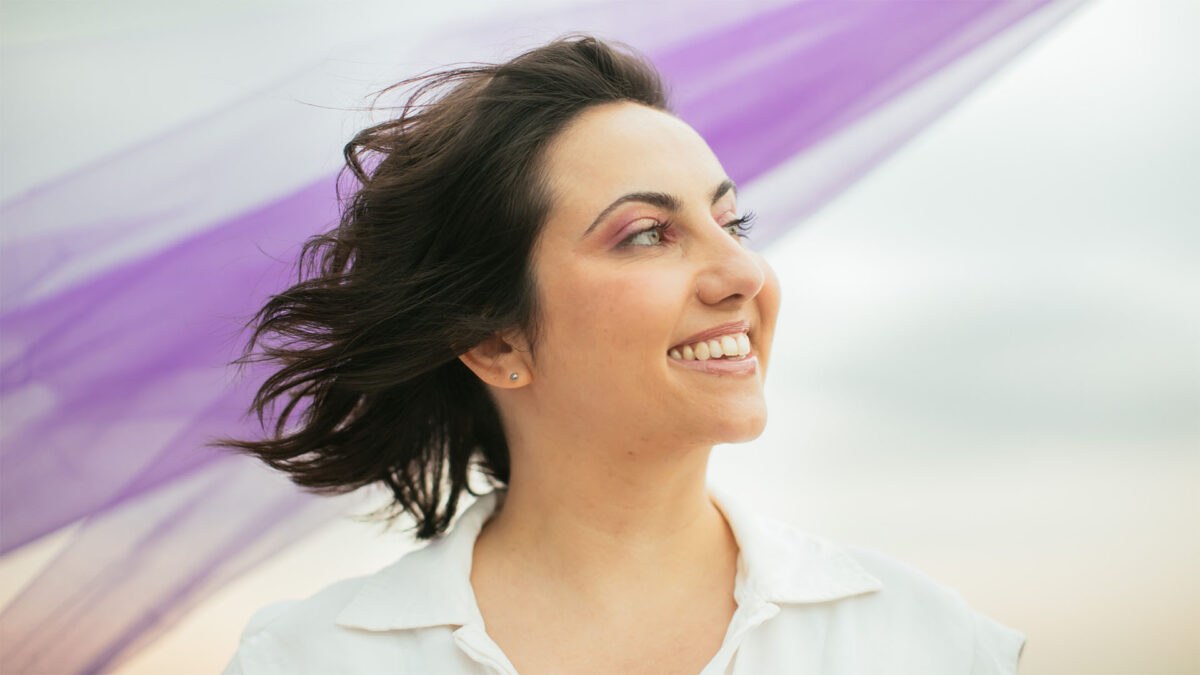 Natie olhando para o lado vestida de branco com tecido roxo ao fundo