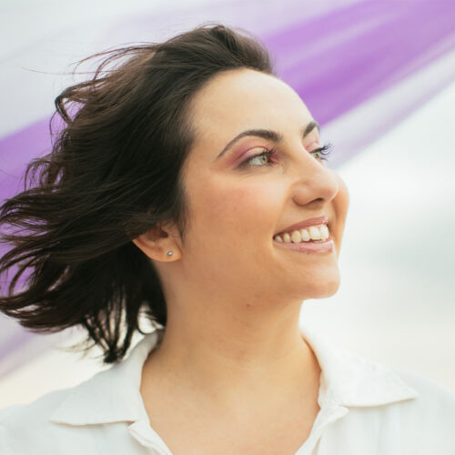 Natie olhando para o lado vestida de branco com tecido roxo ao fundo