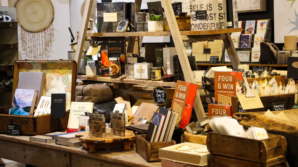 Mesa com prateleira no fundo com grande variedade de produtos, livros, cadernos, adesivos e garrafas