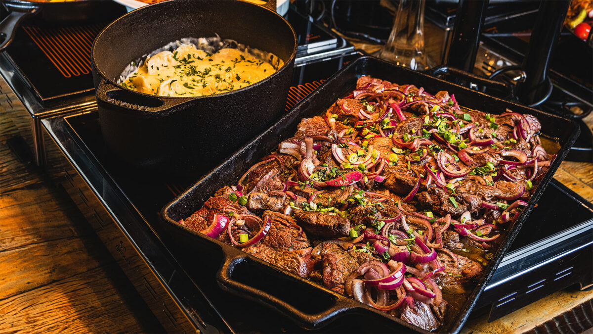 Uma mesa com aquecedor que apoia uma travessa com carnes vermelhas cozidas e uma panela com batatas