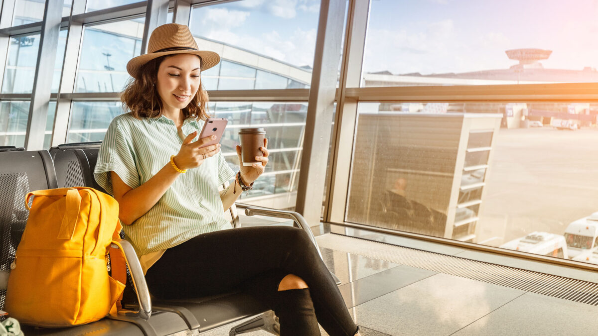 mulher sentada em uma sala de espera de aeroporto enquanto usa o celular com uma mão e segura um copo de café com a outra